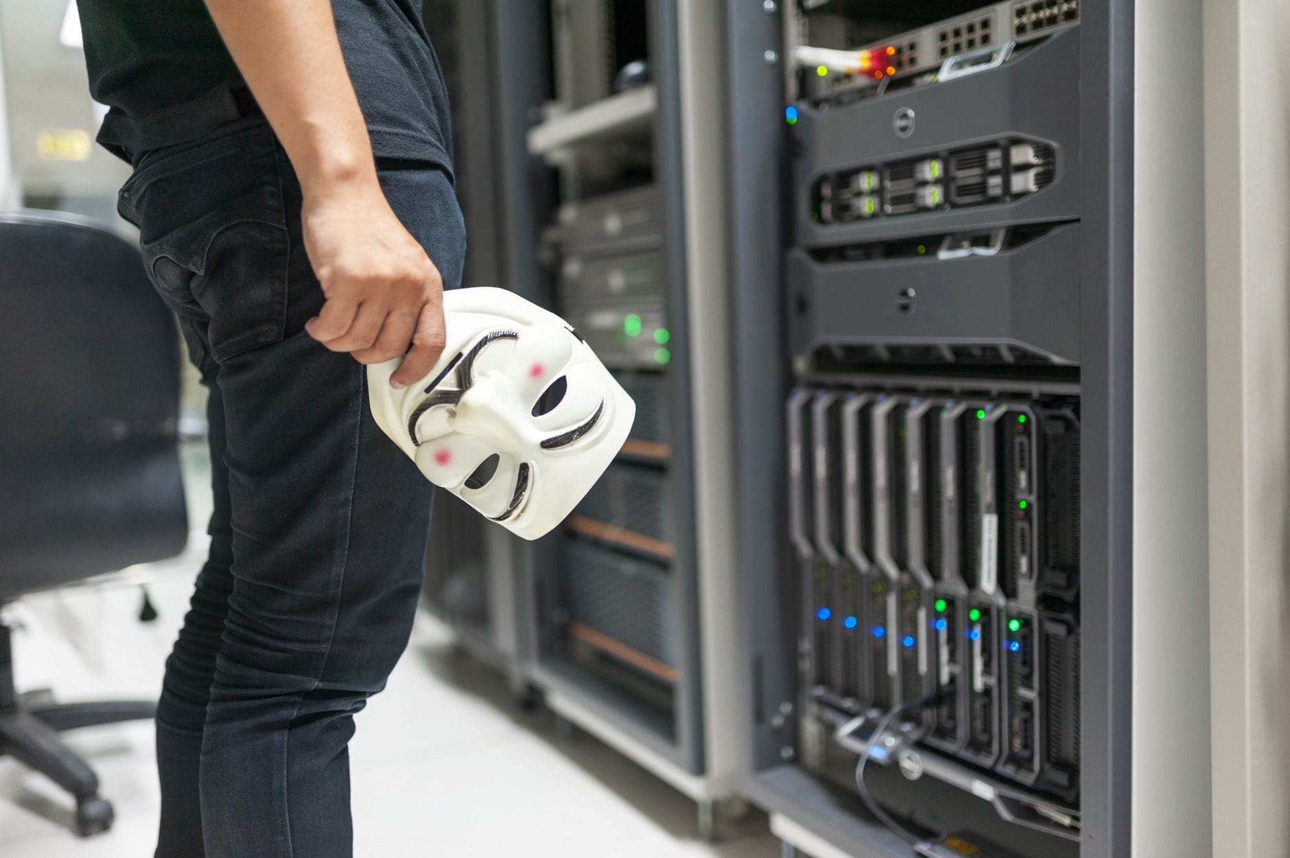 Osoba trzyma maskę anonima na tle serwerów, motyw cyberprzestępczości